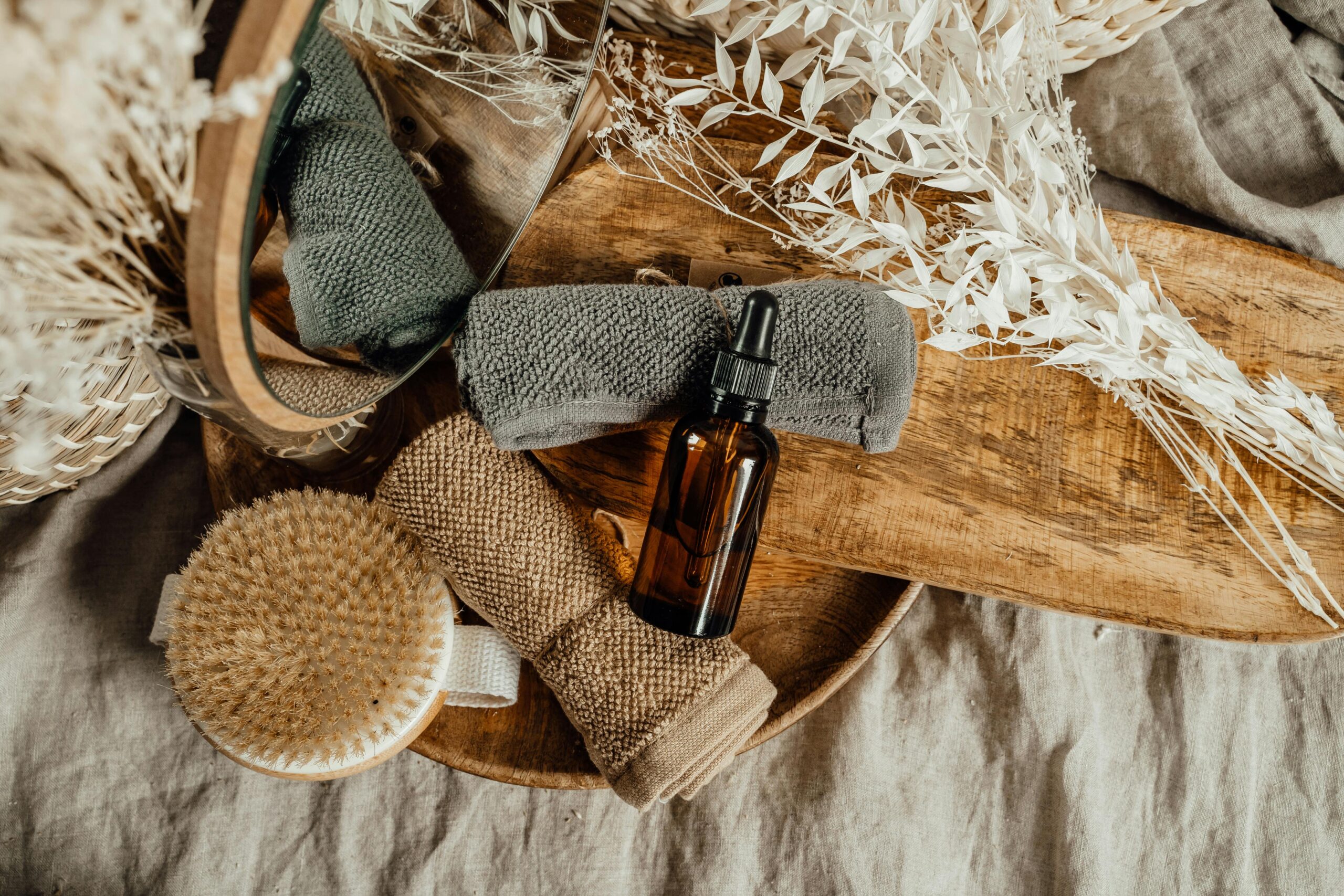 Natural self-care accessories including a glass dropper bottle, exfoliating body brushes, and soft towels displayed on a rustic wooden tray.